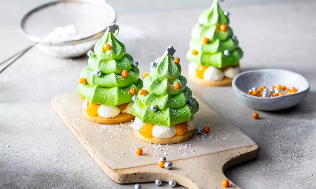 Малки зелени меренг елхички с нежно лъскава текстура върху бисквитена основа, украсени с цветни перли и звездички върху дървена дъска