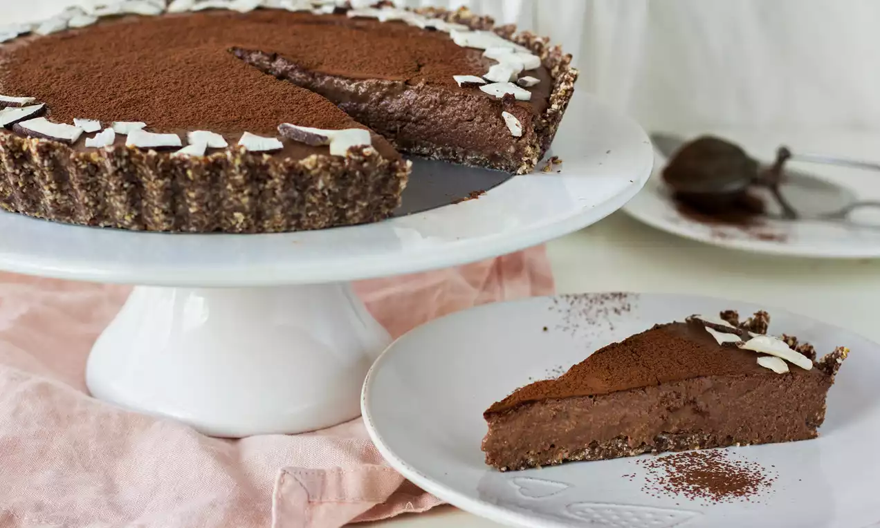 Čokoládovo-kokosový tart s hladkým tmavým krémem, posypaný kakaem a zdobený plátky kokosu, na bílém podnosu a talíři s růžovým ubrusem