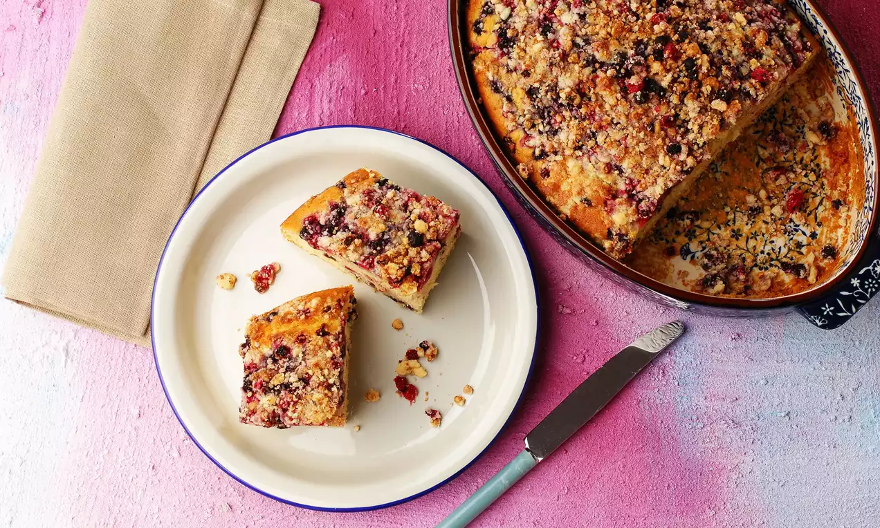 Pembe zeminde beyaz tabakta servis edilen yumuşak dokulu orman meyveli crumble kek, mavi çerçeveli porsiyon ve kırıntılarla süslenmiş