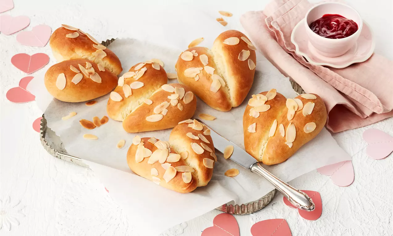 Zlatnosmeđa peciva u obliku srca s prepečenim listićima badema na bijelom papiru, okružena ružičastim srcima i zdjelicom džema