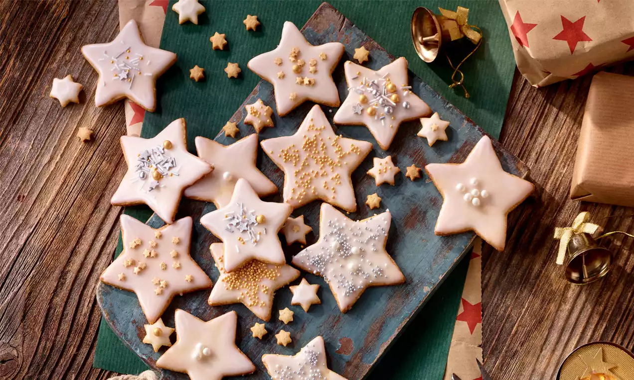 Marzipan-Sternkekse mit weißem Zuckerguss, goldenen und silbernen Verzierungen auf rustikalem Holzbrett, umgeben von kleinen Sternen und Geschenken