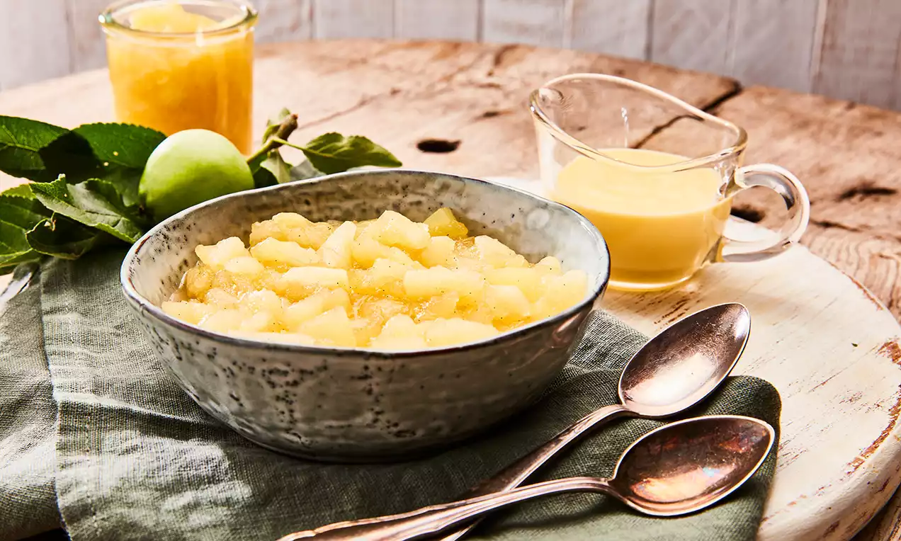 Apró almaszeletek halványsárga kompótban egy szürke kerámiatálban, mellette krémes szósz üvegcsészében és fémkanalak fa tálcán