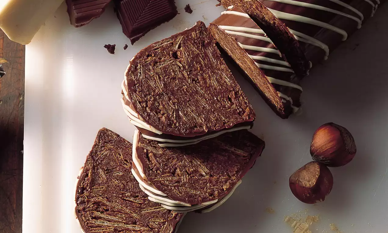 Schokostollen mit dunkler Schokoladenmasse, weißen feinen Streifen, geschnittenen Scheiben, umgeben von Haselnüssen und Waffeln auf weißer Platte