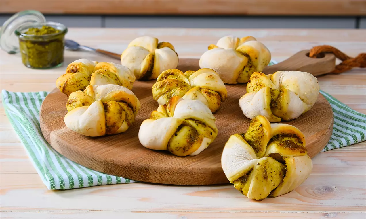 Pestobrötchen mit glänzendem grünem Pesto, goldbrauner Kruste, geschmolzenem Käse und frischen Basilikumblättern auf rustikalem Holzbrett