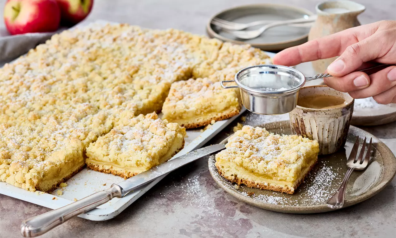 Hand mit Sieb streut Puderzucker auf Apfelmuskuchen mit Hefe vom Blech, saftigem Hefeteig, fruchtiger Apfelmus-Füllung und knusprigen Streuseln – einfaches Kuchenrezept für gemütliche Kaffeerunden.