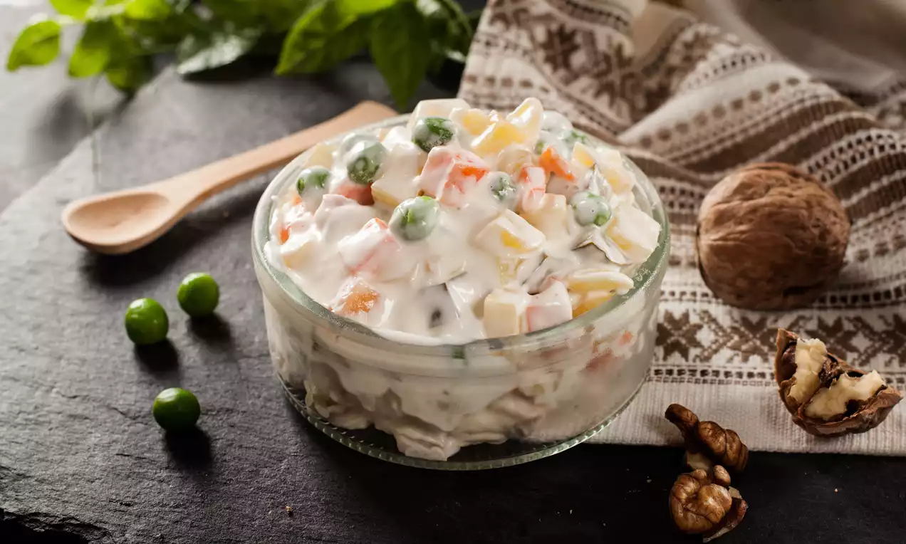 Russian salad in a clear glass bowl with a creamy white dressing, chunks of vegetables and peas, set on a dark slate with walnuts and herbs