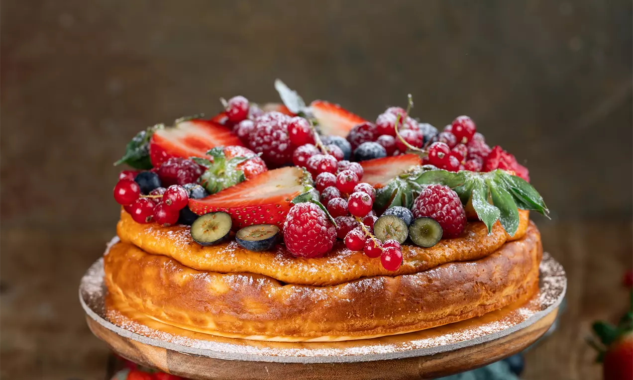 Pečena jogurtova torta z zlato rjavo skorjo, prekrita s svežimi jagodami, rdečim ribezom in borovnicami na lesenem podstavku