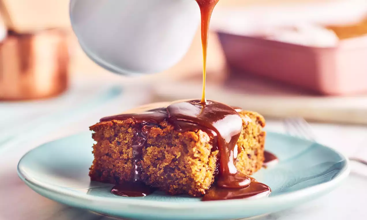 Sticky toffee pudding square with a moist, golden-brown sponge dripping in glossy, amber toffee sauce on a pale blue plate
