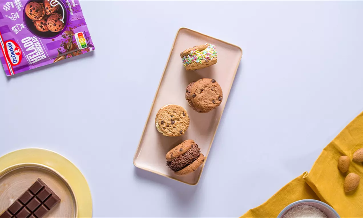 Galletas redondas doradas y marrones rellenas de helado cremoso en tonos pastel sobre bandeja rectangular blanca junto a decoraciones cálidas