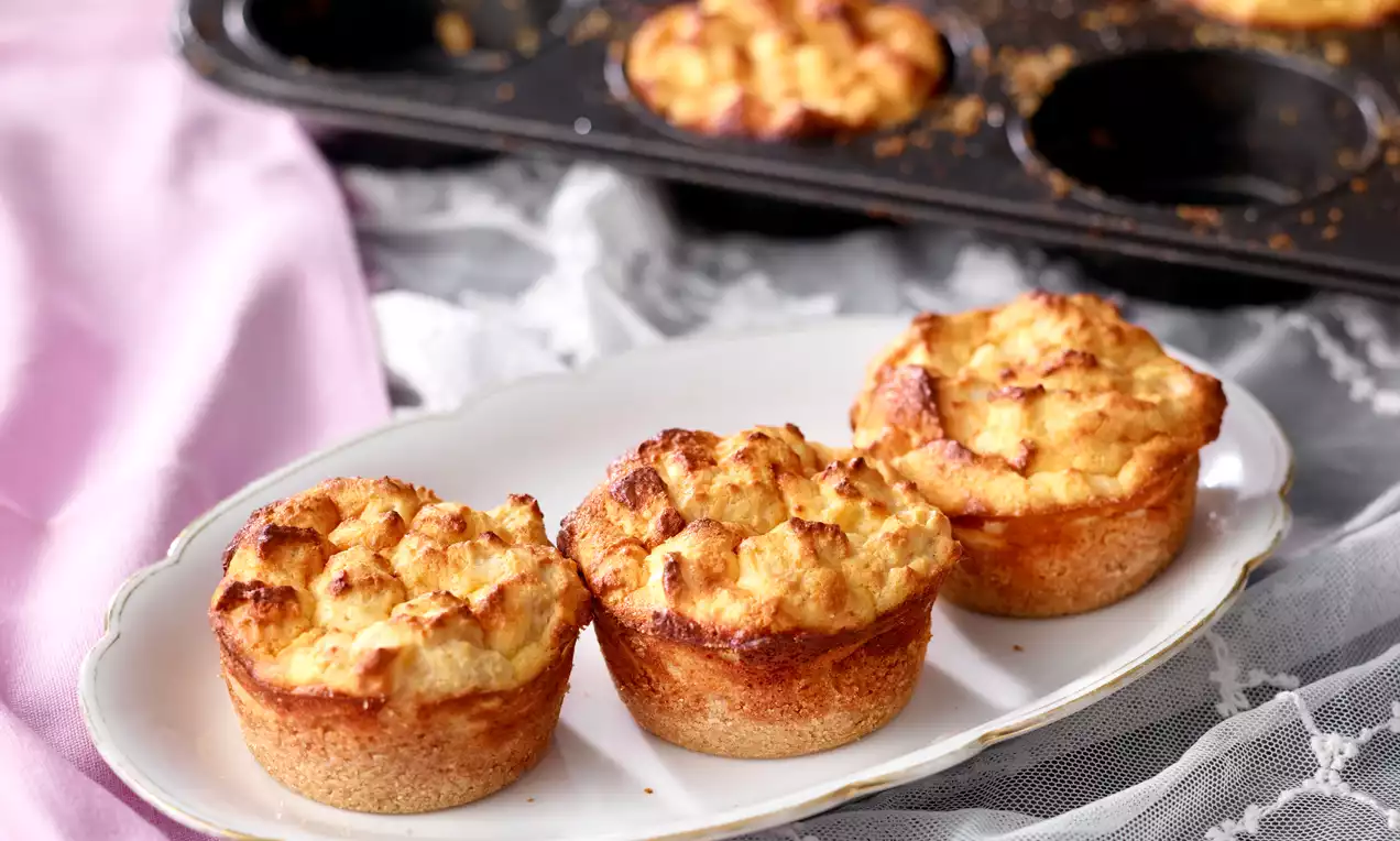 Goldbraune Muffins mit soufflierter, leicht unregelmäßiger Oberfläche auf einem weißen Teller, vor einer schwarzen Backform und einem rosa Tuch drapiert