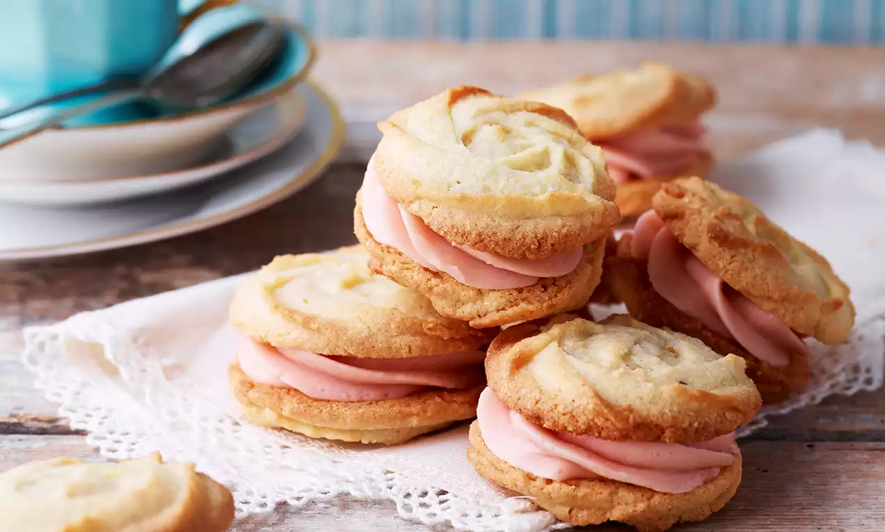 Vaniljeroser med gyldent krumlete småkakepreg og lys rosa hindbærkrem ligger på hvit blonde-serviett på rustikt bord