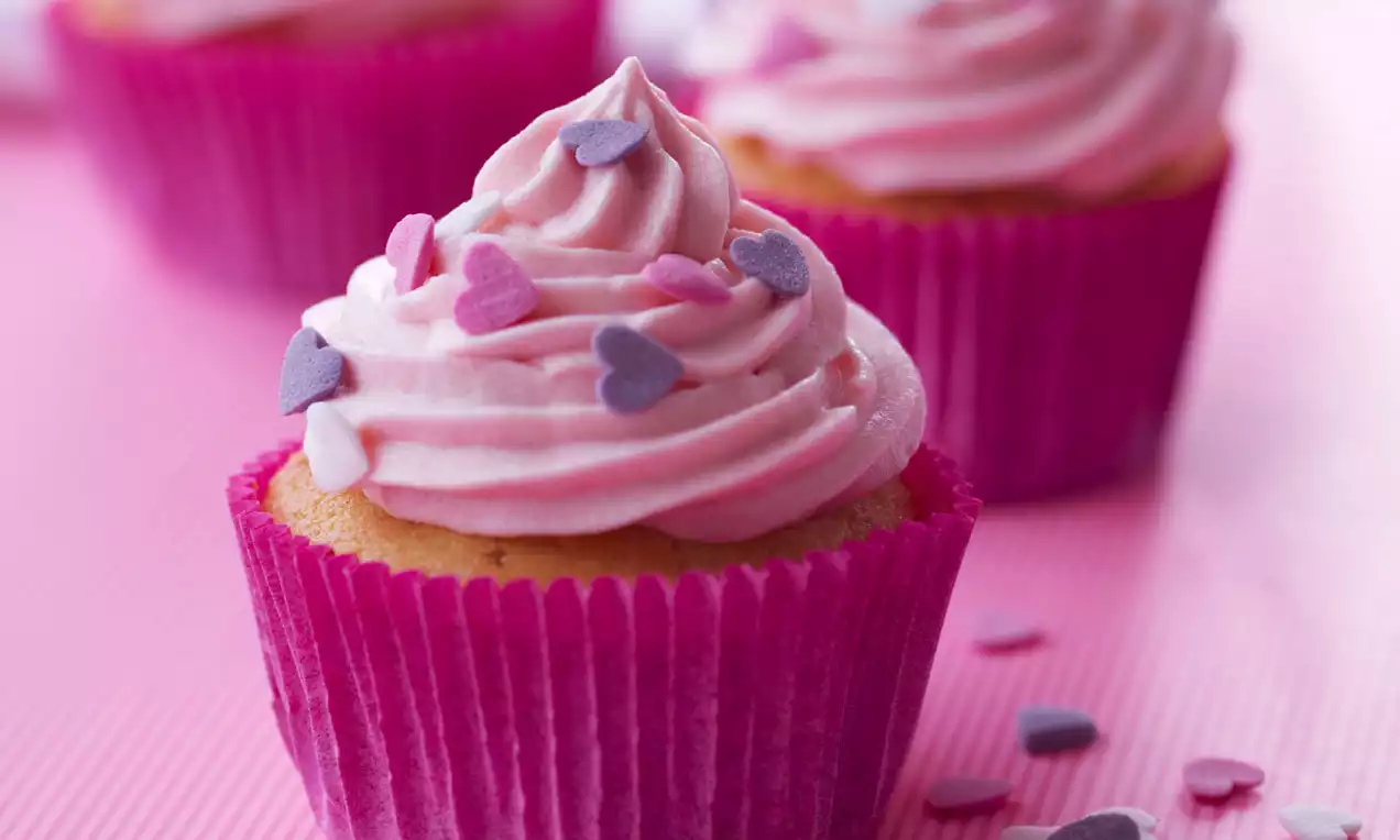 Vaaleanpunaiset cupcaket koristeellisella kermavaahdolla ja sydänkoristeilla, aseteltuna värikkäisiin paperivuokiin vaaleanpunaisella alustalla