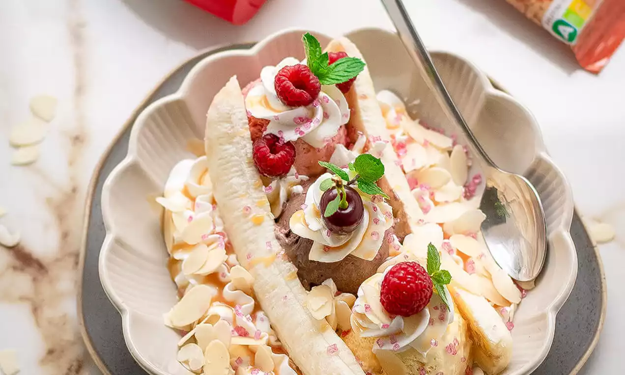 Ledena sladica z rumenimi polovicami banane, kepicami kremastega sladoleda, svežimi malinami, mandljevimi lističi in smetanovimi rozetami na keramičnem krožniku