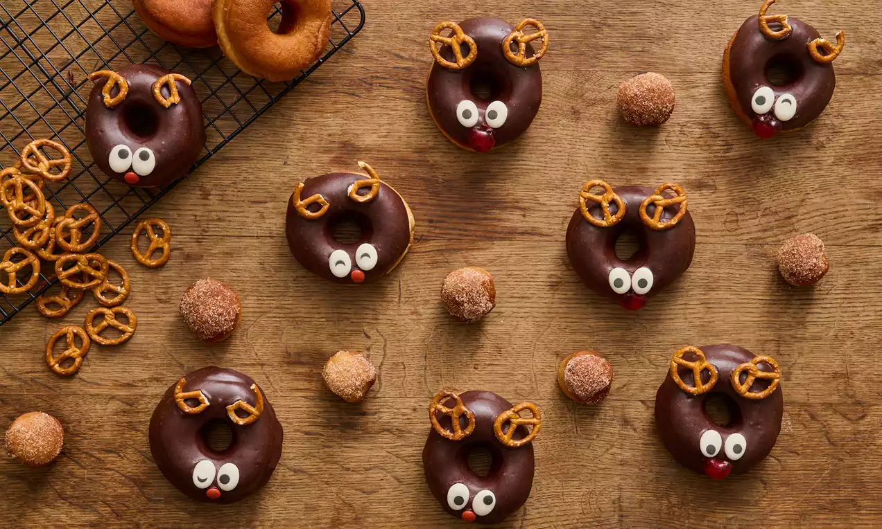 Sjokoladeglasurdekte donuts formet som reinsdyr med hvite øyne, røde neser og saltkjeks-gevir på trebord ved siden av bakverk og gitterrist