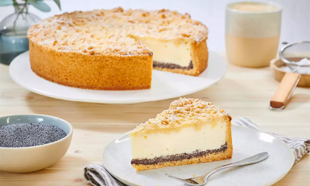 Mohnkuchen mit goldbraunem Streuseldeckel, heller Schmandschicht und dunkler Mohnfüllung auf einem Teller neben Mohnschale und Tasse