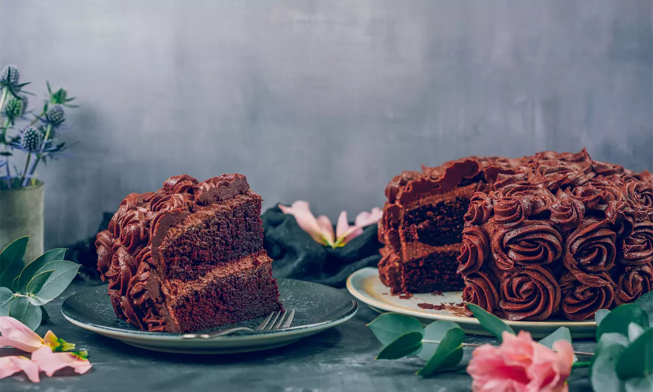 Дълбококафява двуетажна торта с плътен крем, украсена с нежни шоколадови рози върху светла чиния сред зелени листа и розови цветя