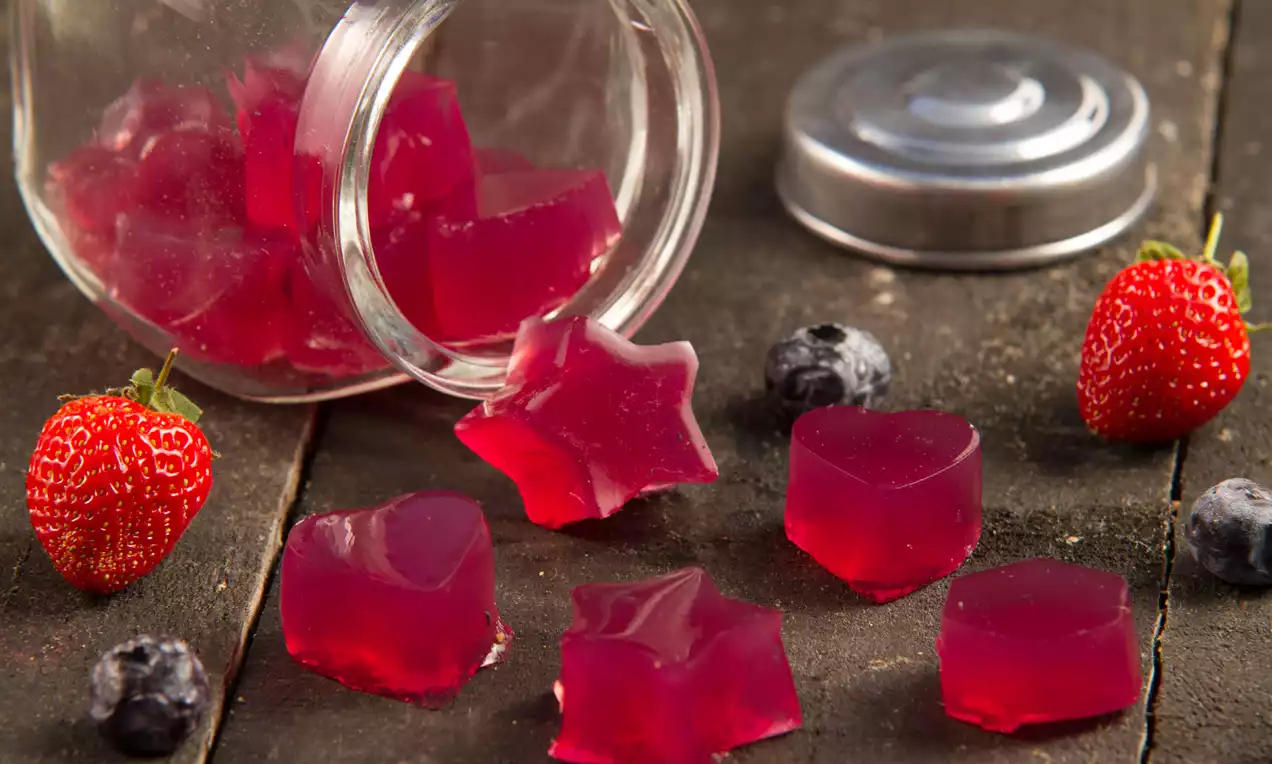 Gelatinas translúcidas de tom vermelho brilhante em formato de estrelas e cubos espalhadas sobre madeira escura ao lado de morangos e blueberries
