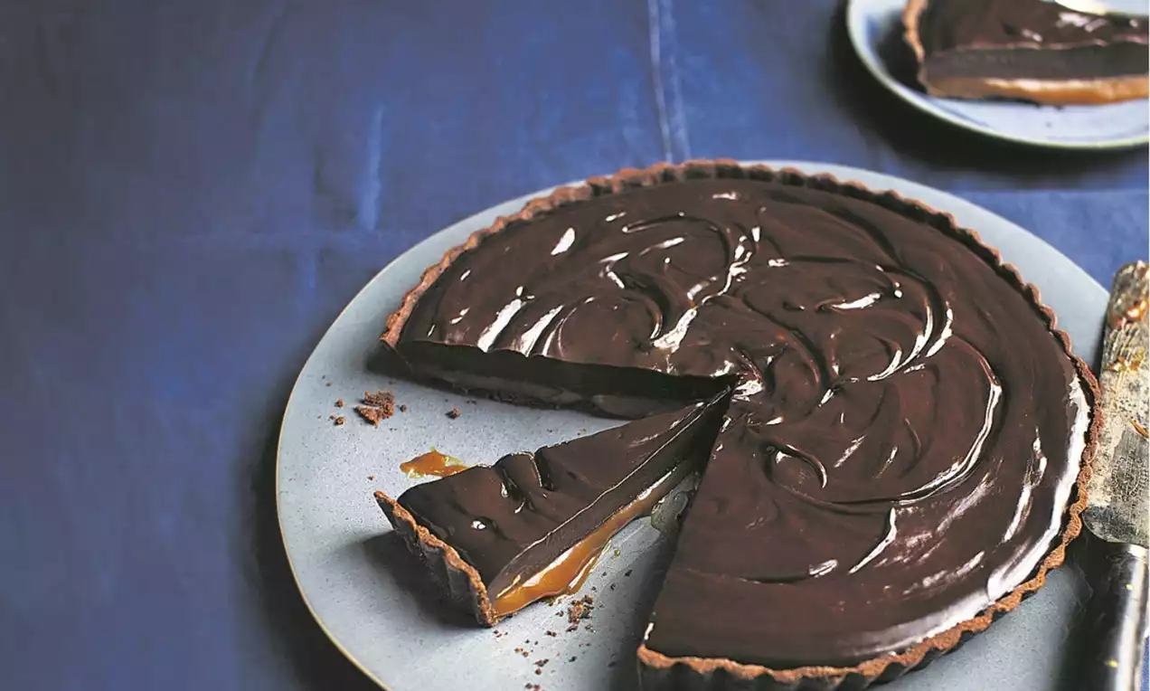 A glossy dark chocolate tart with swirled topping, thin crust, and golden caramel layer, served on a grey plate with one slice cut out