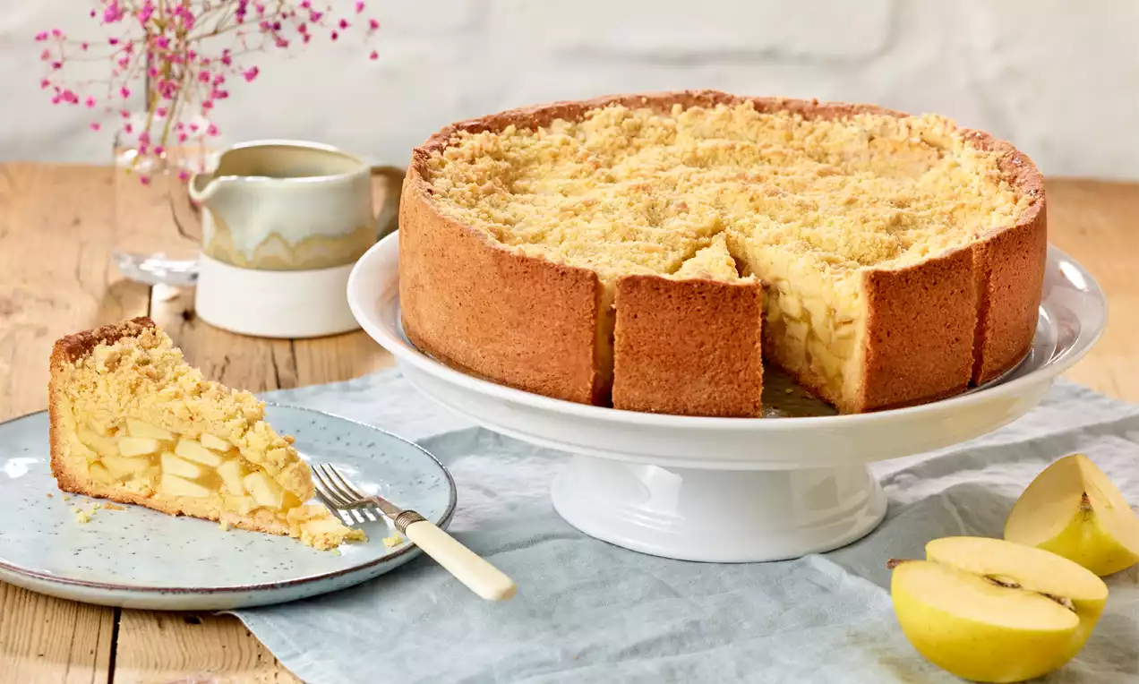 Apfel-Streusel-Torte mit goldbrauner Kruste und hellen Streuseln auf weißer Tortenplatte, daneben ein Stück auf Teller mit Apfelscheiben