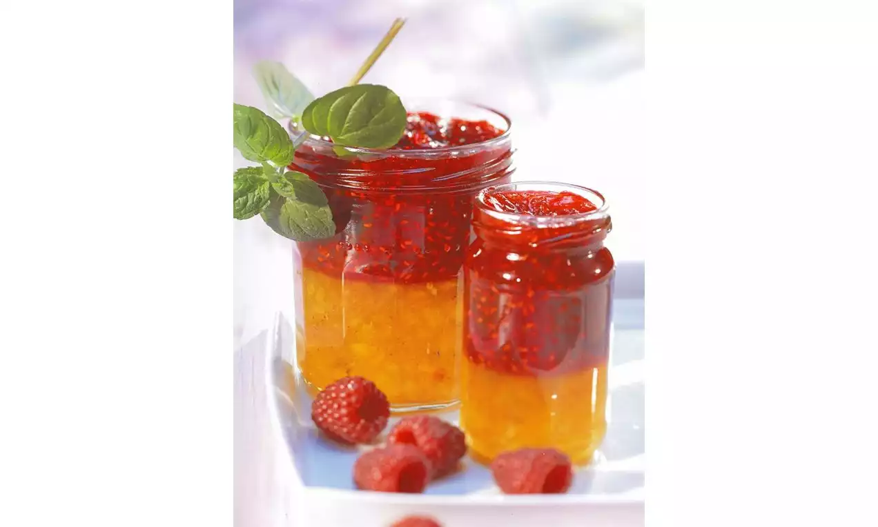 Twee glazen potjes met laagjes goudgele nectarinekleurige en robijnrode frambozenjam, omlijst door verse frambozen en een takje munt