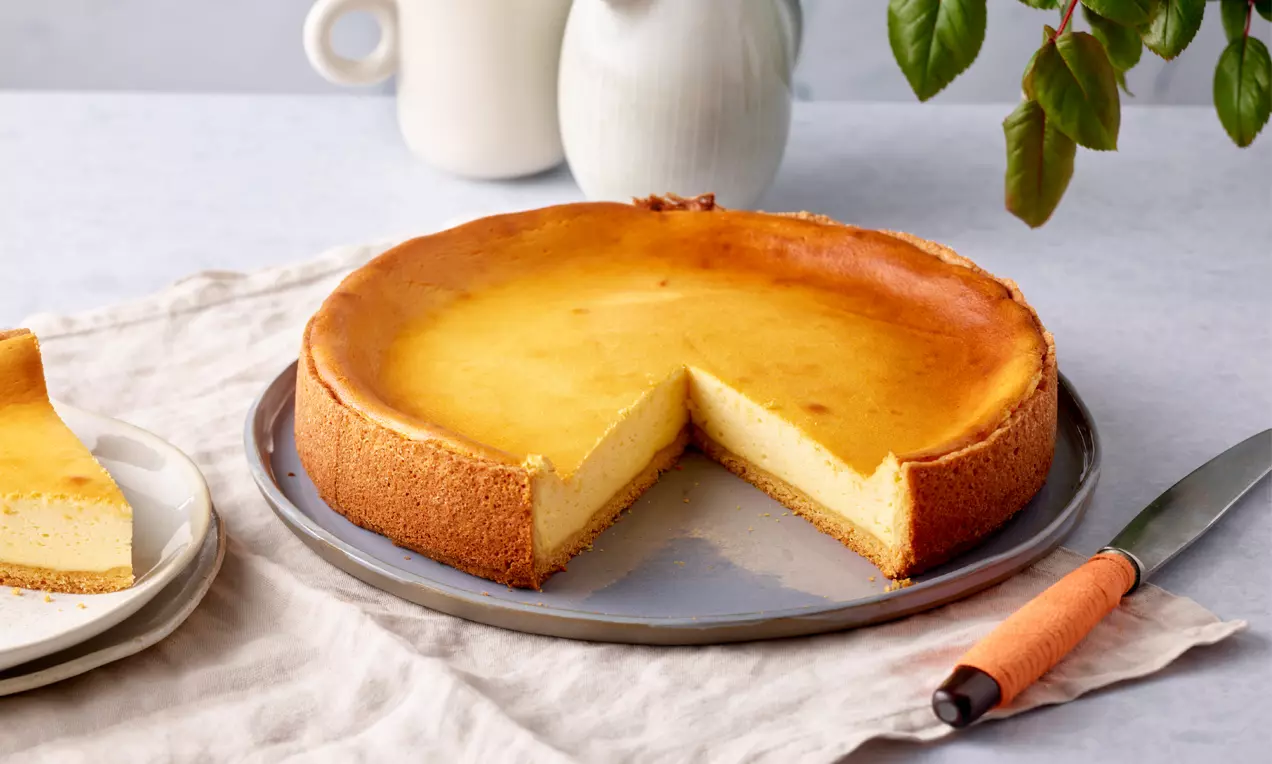 Goldbraune Topfentorte mit samtiger Füllung und glatter Oberfläche, angeschnitten auf metallener Platte neben Teller und Messer