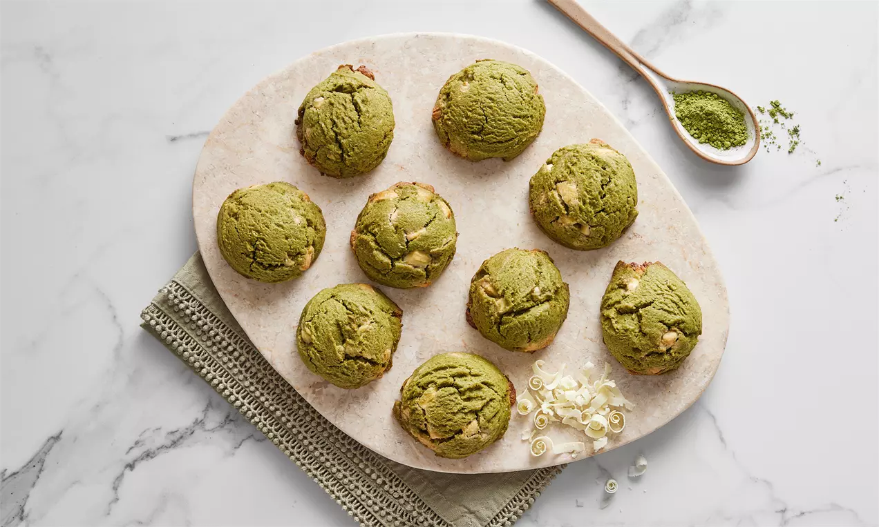 Oval mermer tabakta dizilmiş yuvarlak şekilli, kıvamlı yeşil matcha kurabiyeler, yanında matcha tozu ve beyaz çikolata parçaları eşlik ediyor