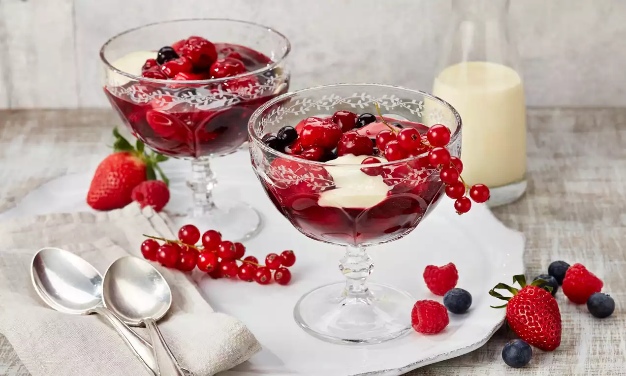 Rote Grütze in gläsernen Dessertschalen mit roten und dunklen Beeren, hellgelben Sahneflecken, daneben frische Beeren und Sahne im Hintergrund