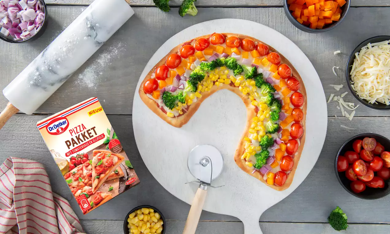 Regenboogvormige pizza op een houten bord met laagjes helderrode tomaat, groene broccoli, gele maïs en paarse ui tussen kaasschilfers