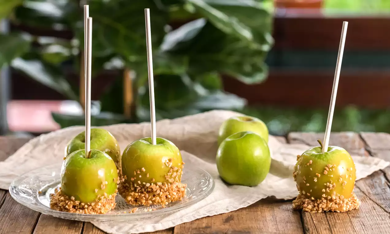 Manzanas verdes brillantes cubiertas de caramelo dorado y trozos de almendra, con palitos plateados sobre madera y un fondo verde