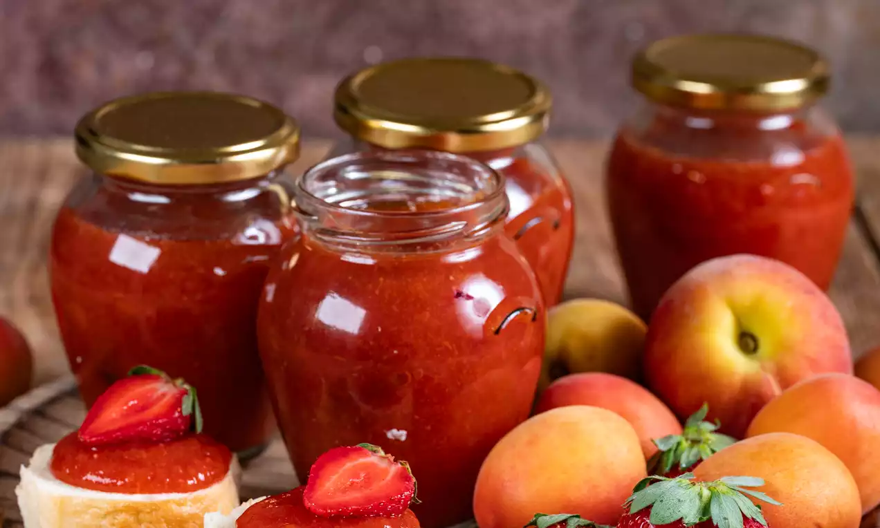 Staklenke miješanog pekmeza crvenkasto-narančaste boje, okružene svježim marelicama i jagodama na drvenom tanjuru