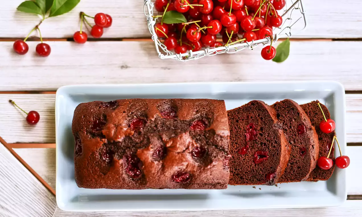 Podłużna babka czekoladowa na białej paterze, ciemnobrązowa z soczystymi wiśniami w cieście i dekoracją z gałązkami świeżych wiśni