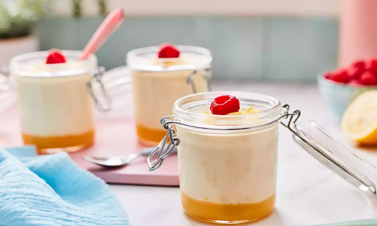 Creamy pale lemon mousse layered over glossy golden sauce in glass jars with hinged lids, topped with a single bright red raspberry