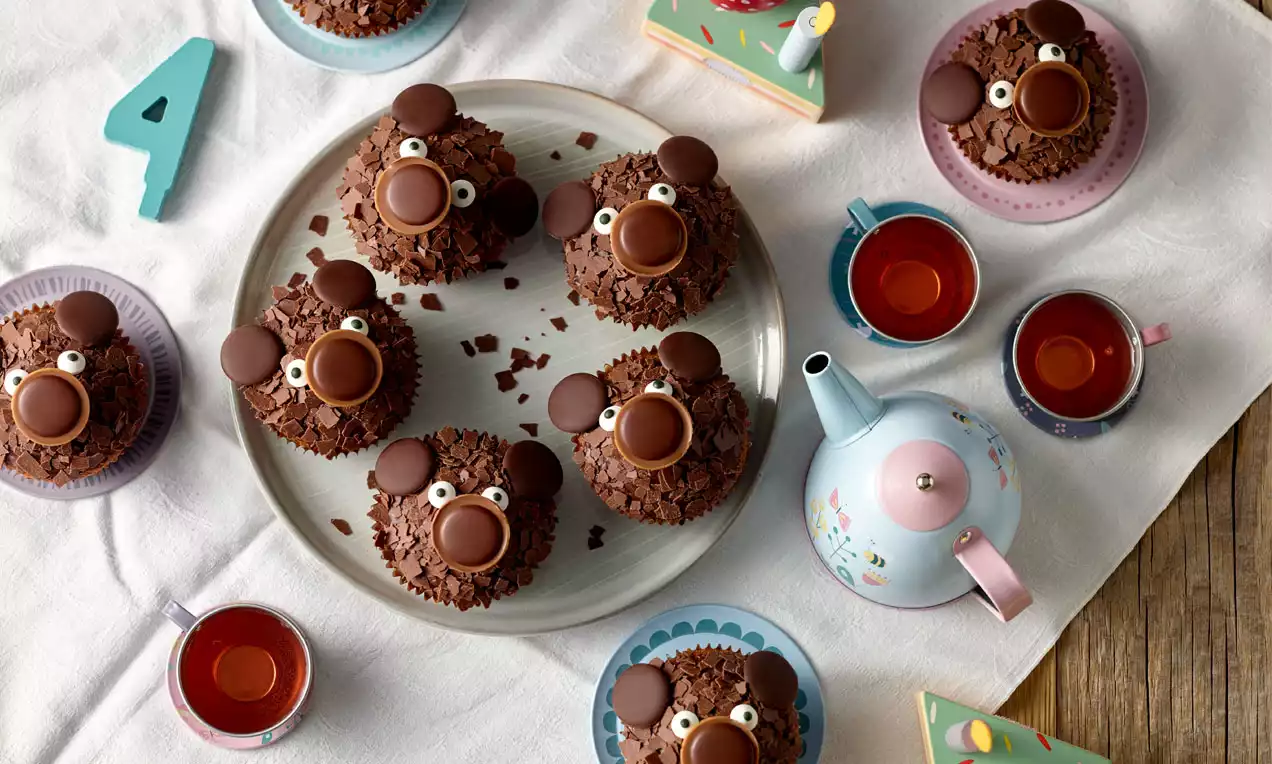 Schokoladen-Muffins mit braunen Ohren, runder Nase und weißen Augen auf einem Teller, umgeben von bunten Tassen und Teekanne