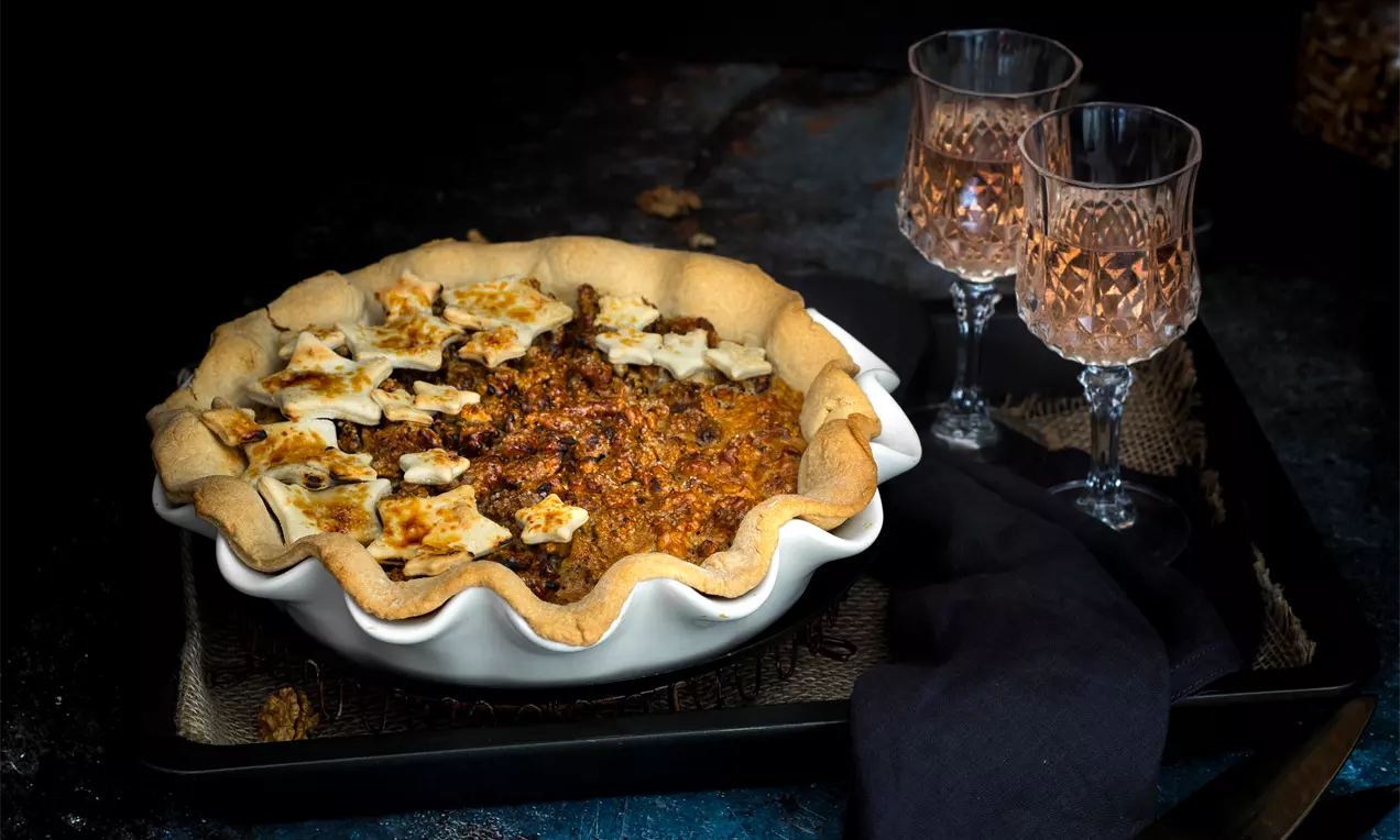 Кафяв орехов пай със златиста коричка, декориран с маслени тестени звездички, разположен в бяла чиния върху тъмна повърхност