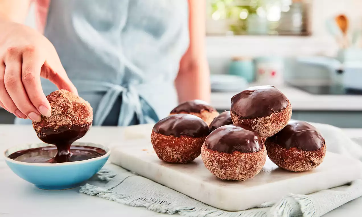 Golden brown donuts coated in sugar, some topped with glossy chocolate glaze, arranged on a marble slab with one being dipped in a bowl of chocolate