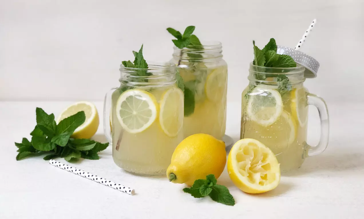 Светложълта лимонада в стъклени буркани с резени лимон, свежи ментови листа и хартиени сламки сред цели и разполовени лимони