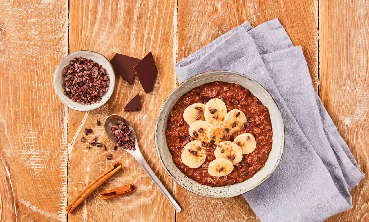 Gusta, tamnosmeđa čokoladna kaša u keramičkoj zdjeli ukrašena svježim kolutovima banane i komadićima kakao zrna na drvenoj podlozi