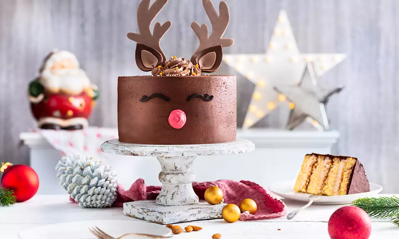 Tarta circular de chocolate decorada con cuernos marrones, ojos y nariz roja sobre base blanca, rodeada de adornos navideños