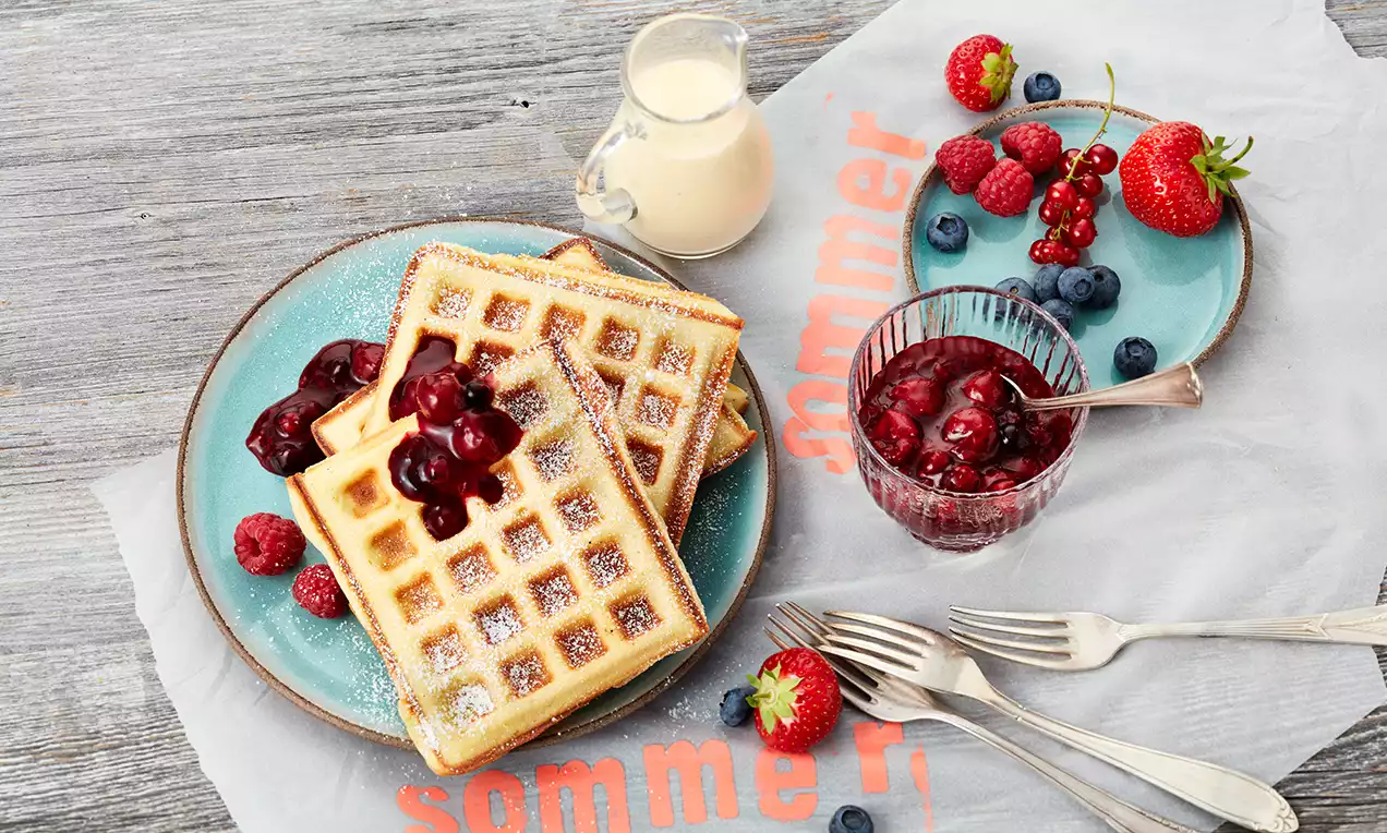 Goldgelbe Waffeln mit feinem Puderzucker bestäubt, gestapelt auf einem türkisfarbenen Teller, garniert mit roten Beeren und Kirschkompott