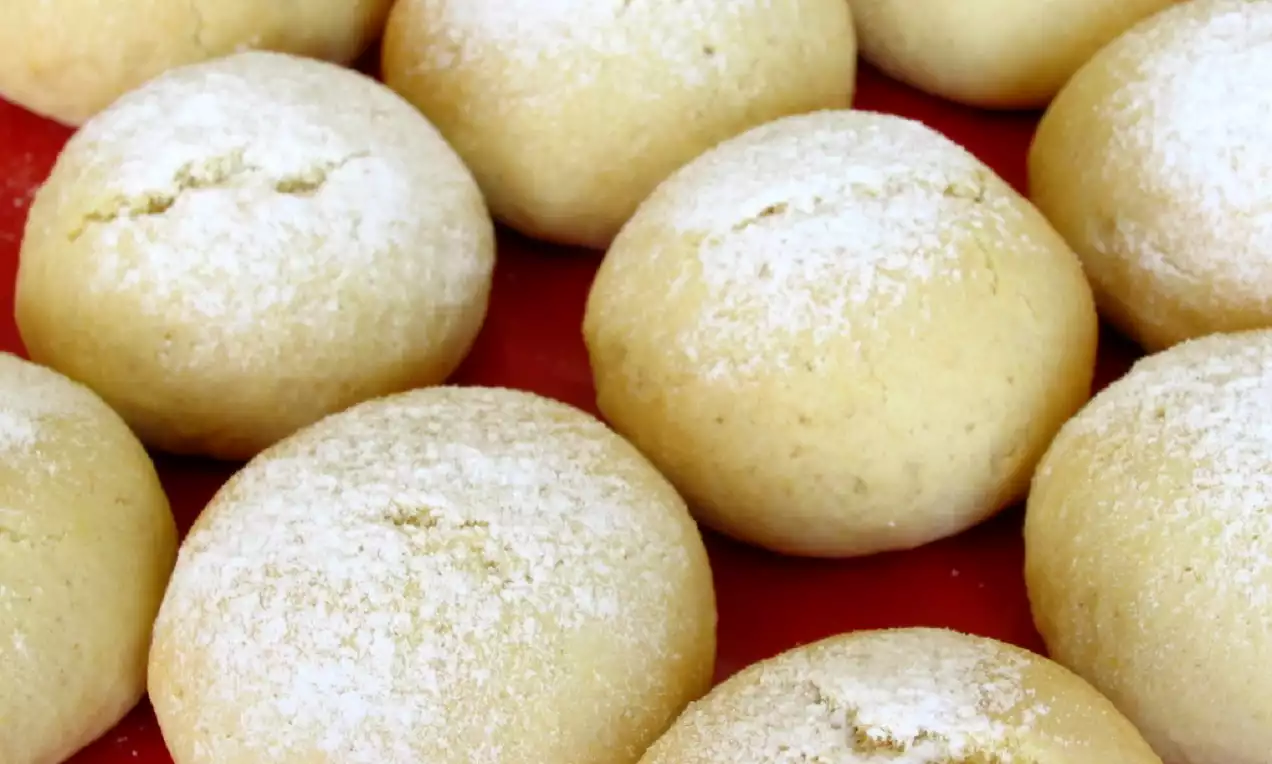 Altın sarısı yuvarlak kurabiyeler, pürüzsüz ve hafif çatlak yüzeyleriyle beyaz pudra şekeri serpilmiş kırmızı zemin üzerine dizilmiş