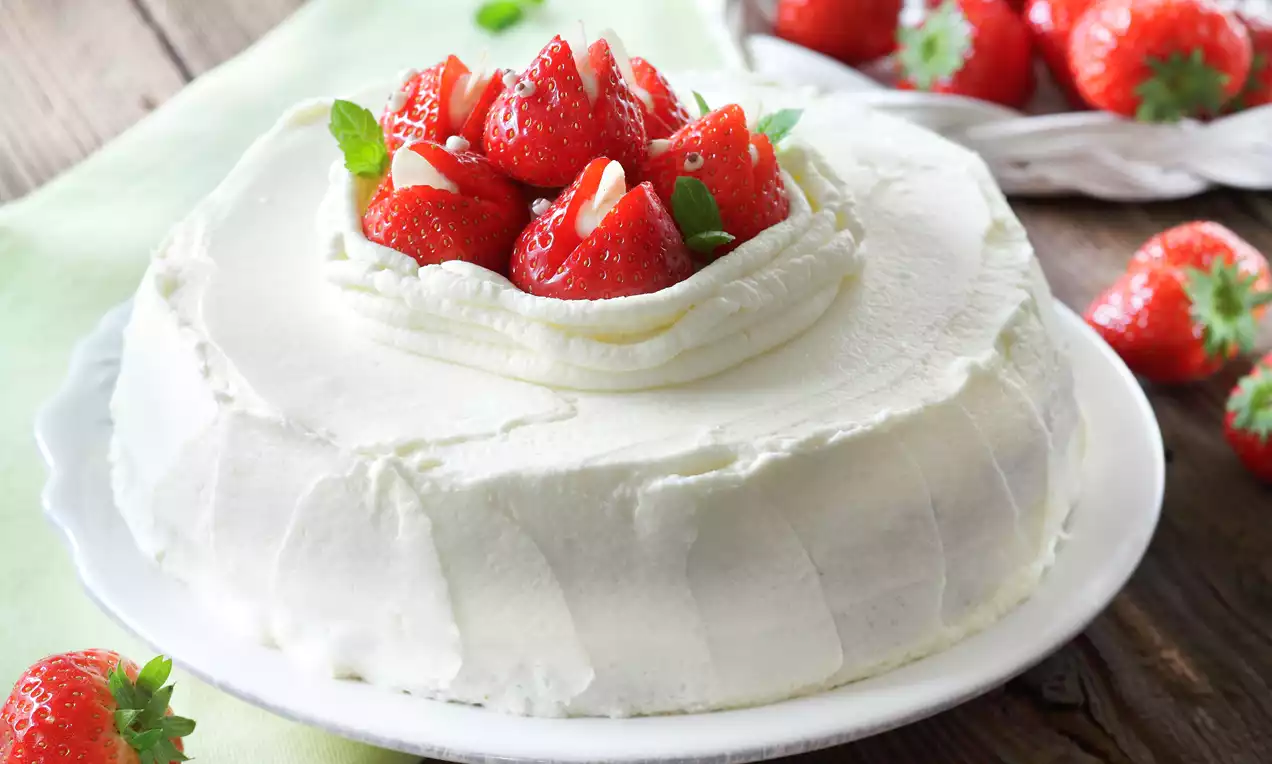 Tort o gładkiej białej kremowej powierzchni, udekorowany świeżymi truskawkami, płatkami migdałów i listkami mięty na białym talerzu