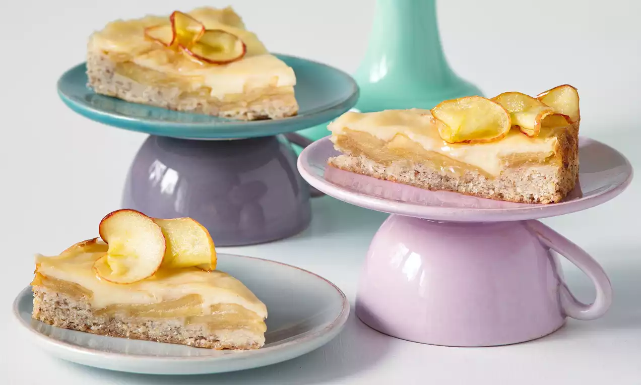 Trojúhelníkové porce světle hnědého koláče s jemnou vrstvou jablek a krémem, zdobené sušenými plátky jablek na pastelových talířích