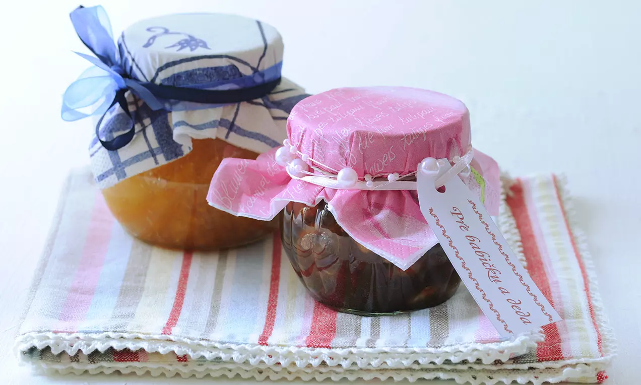 Dva skleněné zavařovací sklenice s barevnými textilními víčky, jedna modrobílá a druhá růžová, položeny na pruhované látce.