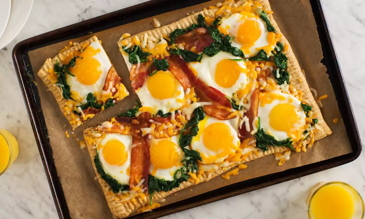 A rectangular breakfast tart with sunny-side-up eggs, crispy bacon, melted cheese, and sautéed spinach on a golden crust-lined tray