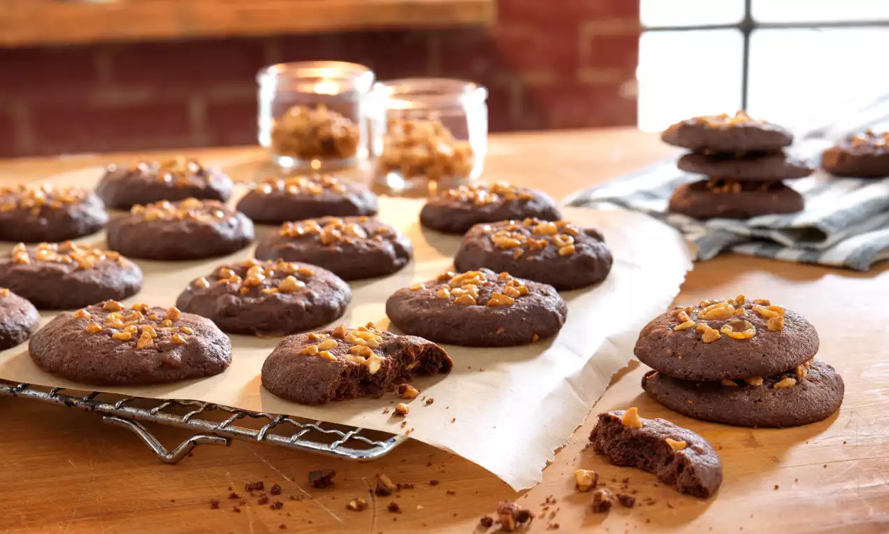Schokoladenbraune Cookies mit goldbraunen Erdnussstücken auf Backpapier und Gitter, neben gestapelten Cookies und Glasbehältern auf Holztisch