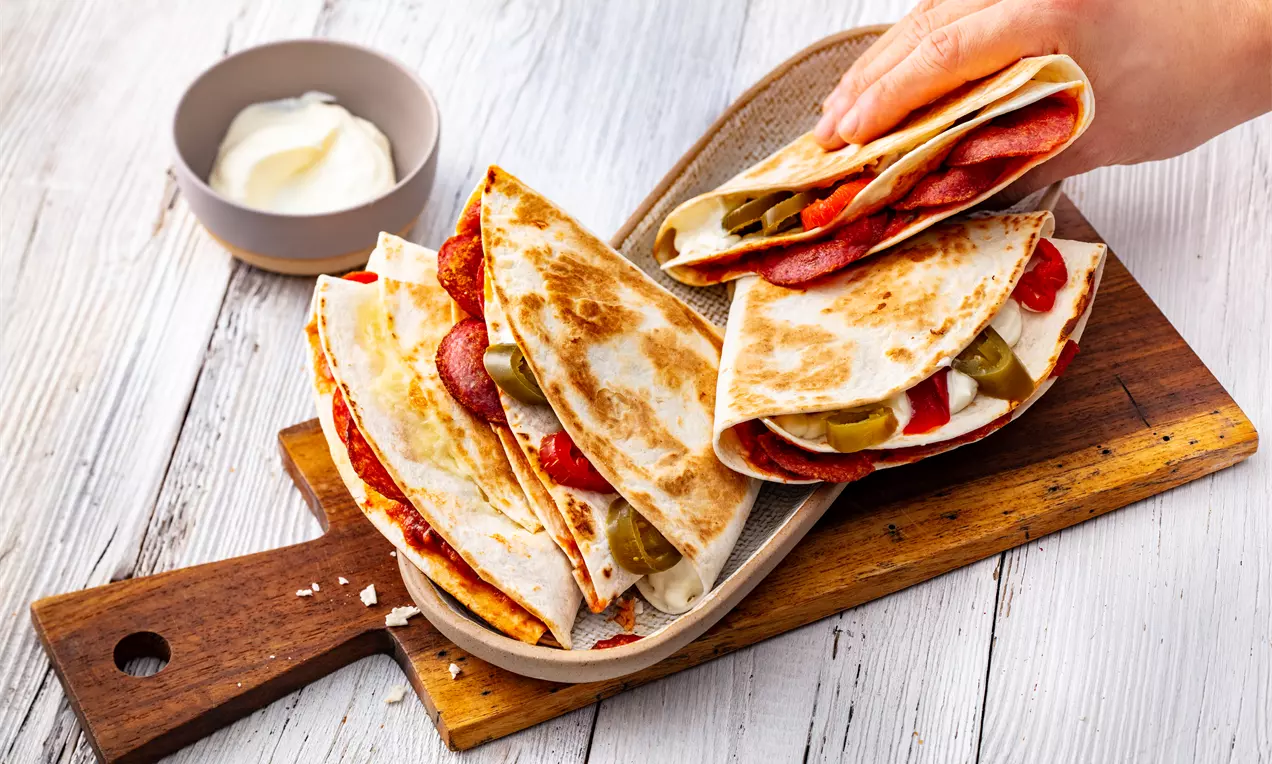 Тортии със златист загар, сгънати на две и пълни с червени колелца салам, зелени чушки и топящо се сирене върху дървена дъска