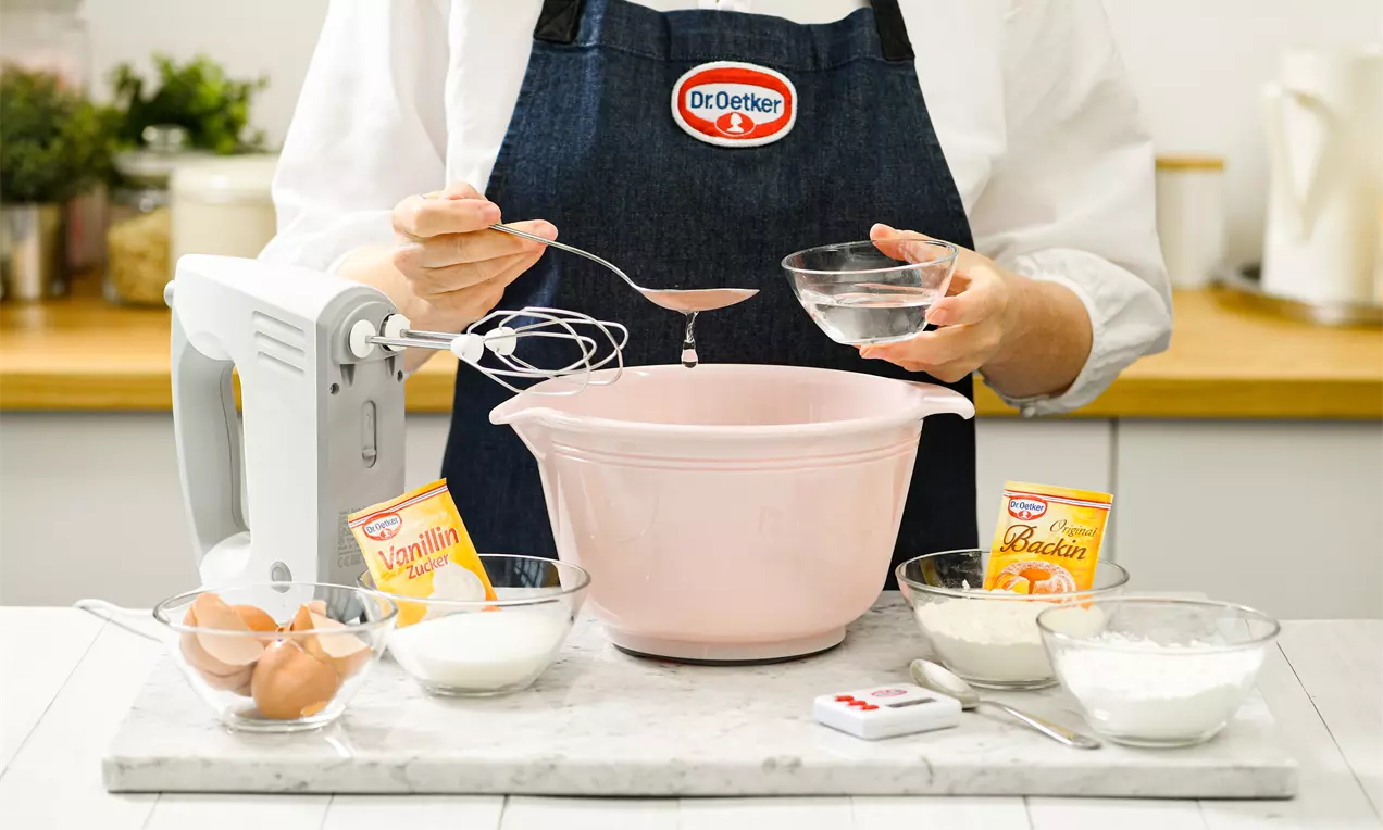 Светлорозова купа с гладка повърхност, пълна със съставки, заобиколена от правилно подредени продукти за печене върху светъл плот