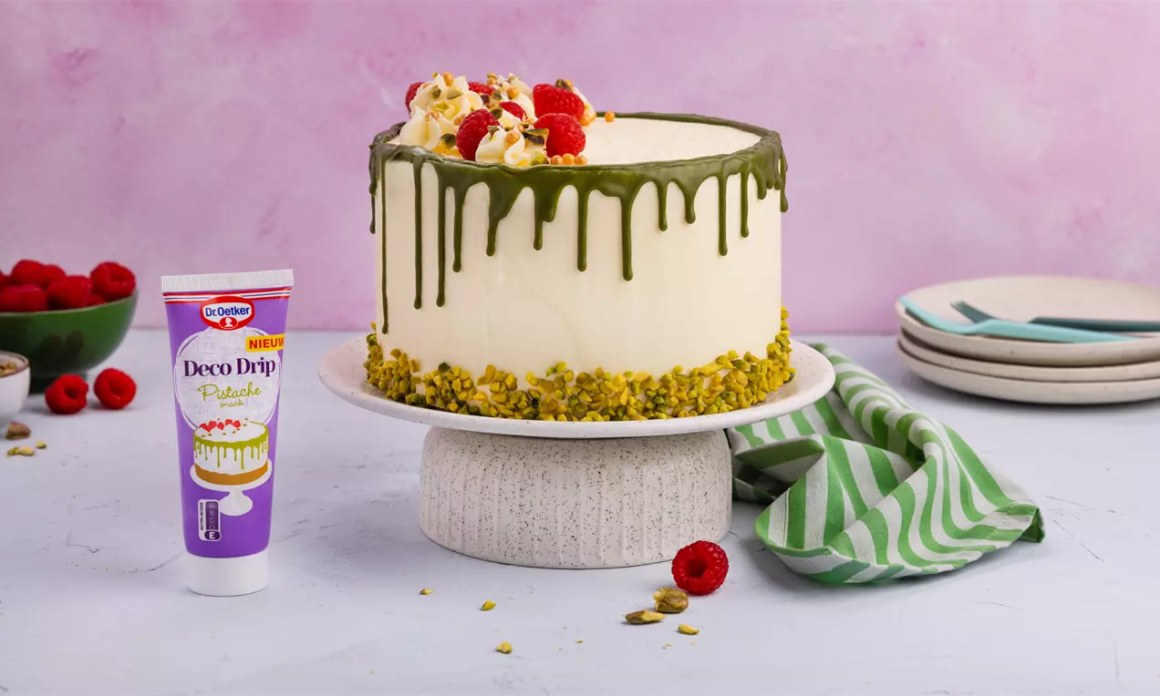 Witte dripcake met groene pistacheglazuur, bestrooid met fijngemalen pistachenoten en versierd met frambozen en gebakdecoratie op een gebroken wit taartplateau