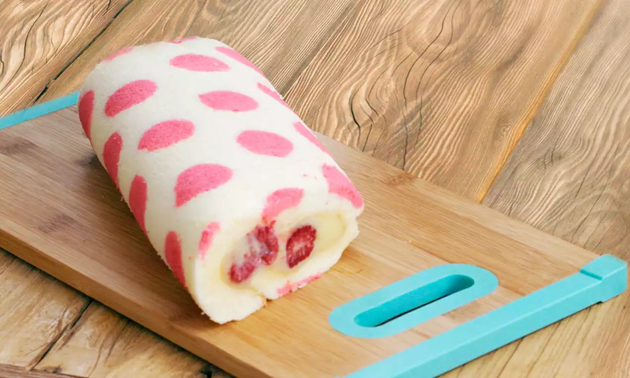 Opgerolde witte cake met roze stippen, gevuld met room en stukjes aardbei, op een houten snijplank met turquoise rand