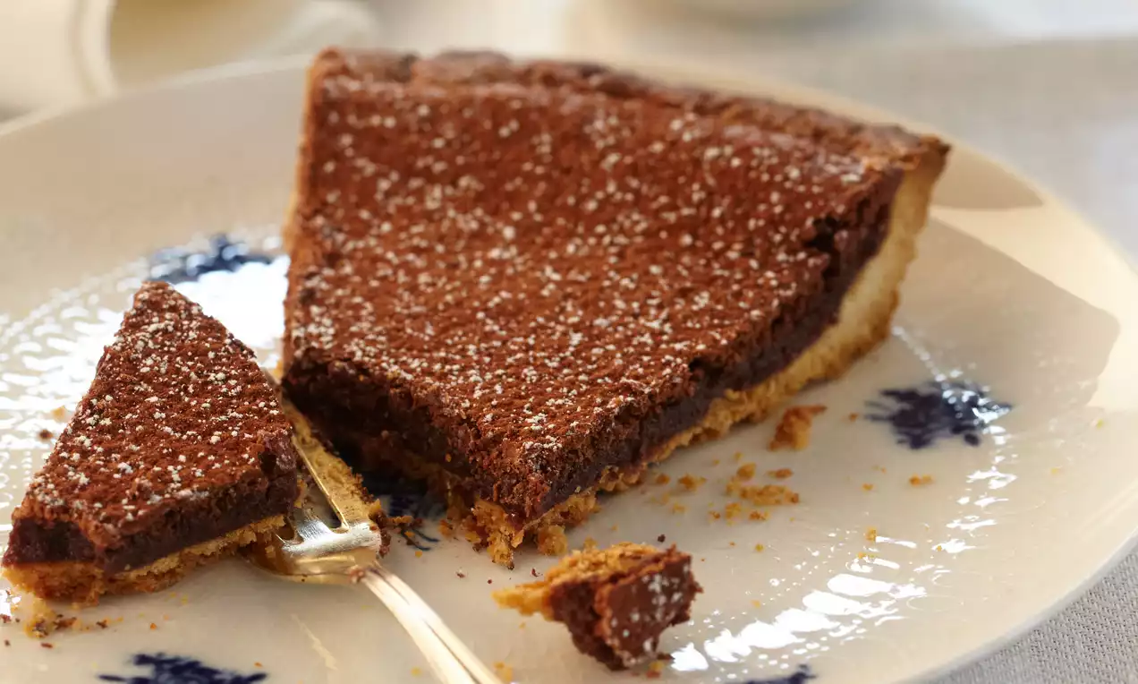 Tarte au chocolat brun foncé sur une pâte dorée, légèrement saupoudrée de sucre glace blanc, présentée sur une assiette en porcelaine blanche et bleue